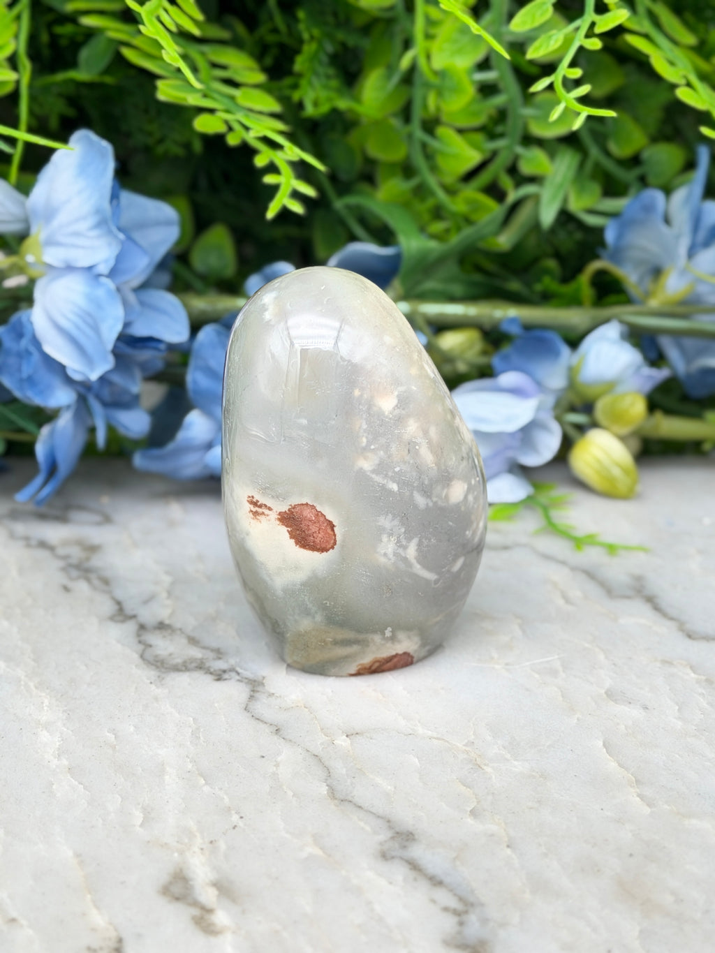 a gray rock with a brown spot on it, placed on a marble surface. The rock is surrounded by blue flowers, creating a visually appealing contrast.