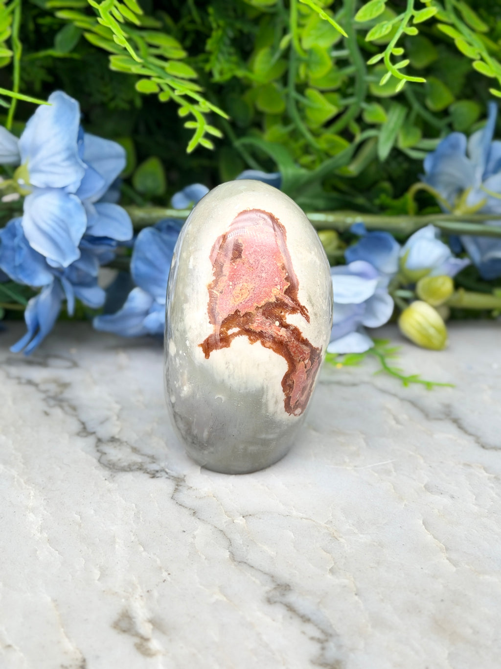 a white rock with a pinkish-brown pattern on it, placed on a marble surface. The rock is surrounded by blue flowers, creating a visually appealing contrast.