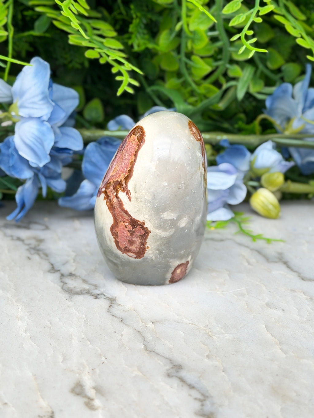 a white and brown speckled rock with a reddish-brown speckled pattern, placed on a marble surface. The rock is surrounded by blue flowers, creating a visually appealing contrast between the natural and man-made elements.