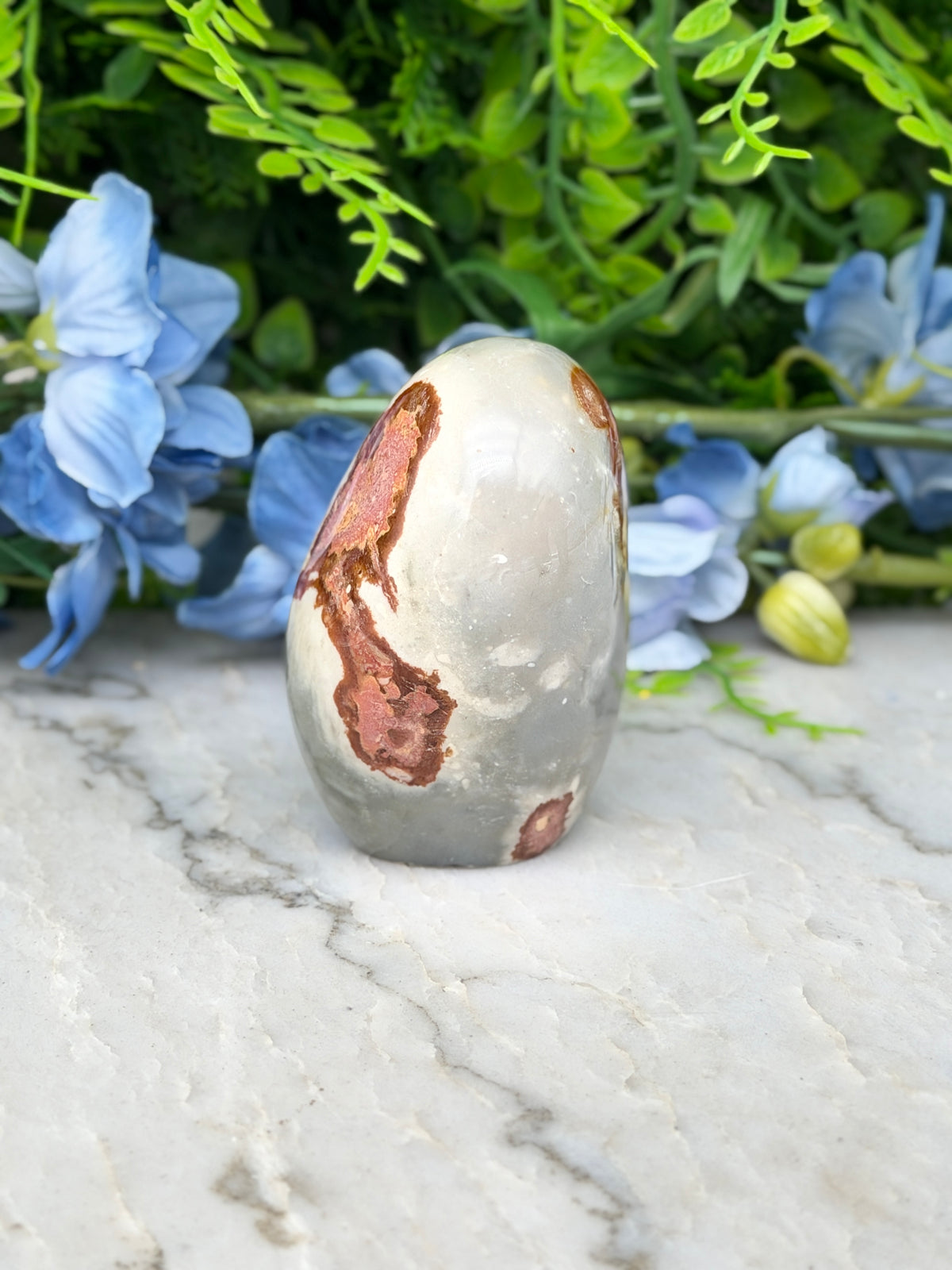 a white and brown speckled rock with a reddish-brown speckled pattern, placed on a marble surface. The rock is surrounded by blue flowers, creating a visually appealing contrast between the natural and man-made elements.
