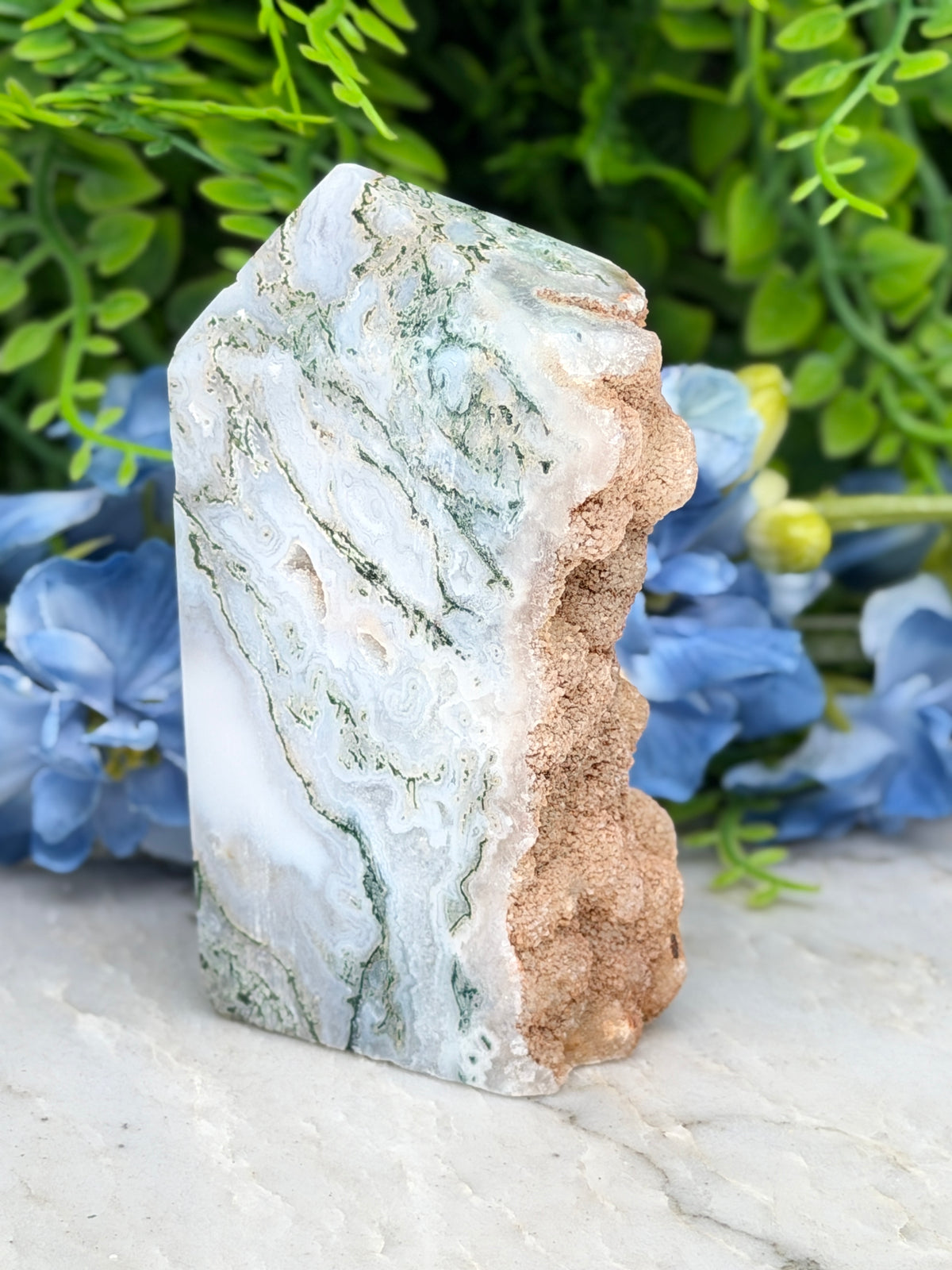 A jadeite crystal with a light green and white striped pattern sits on a white surface, surrounded by blue flowers.