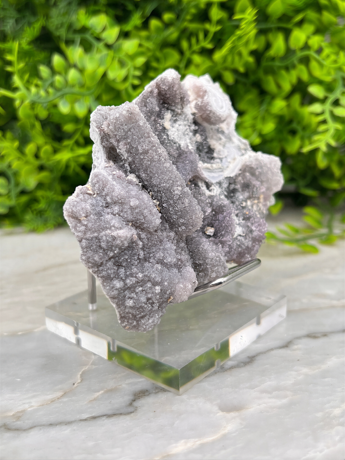 a piece of purple crystal or rock on a clear glass stand, placed on a marble surface with greenery in the background.