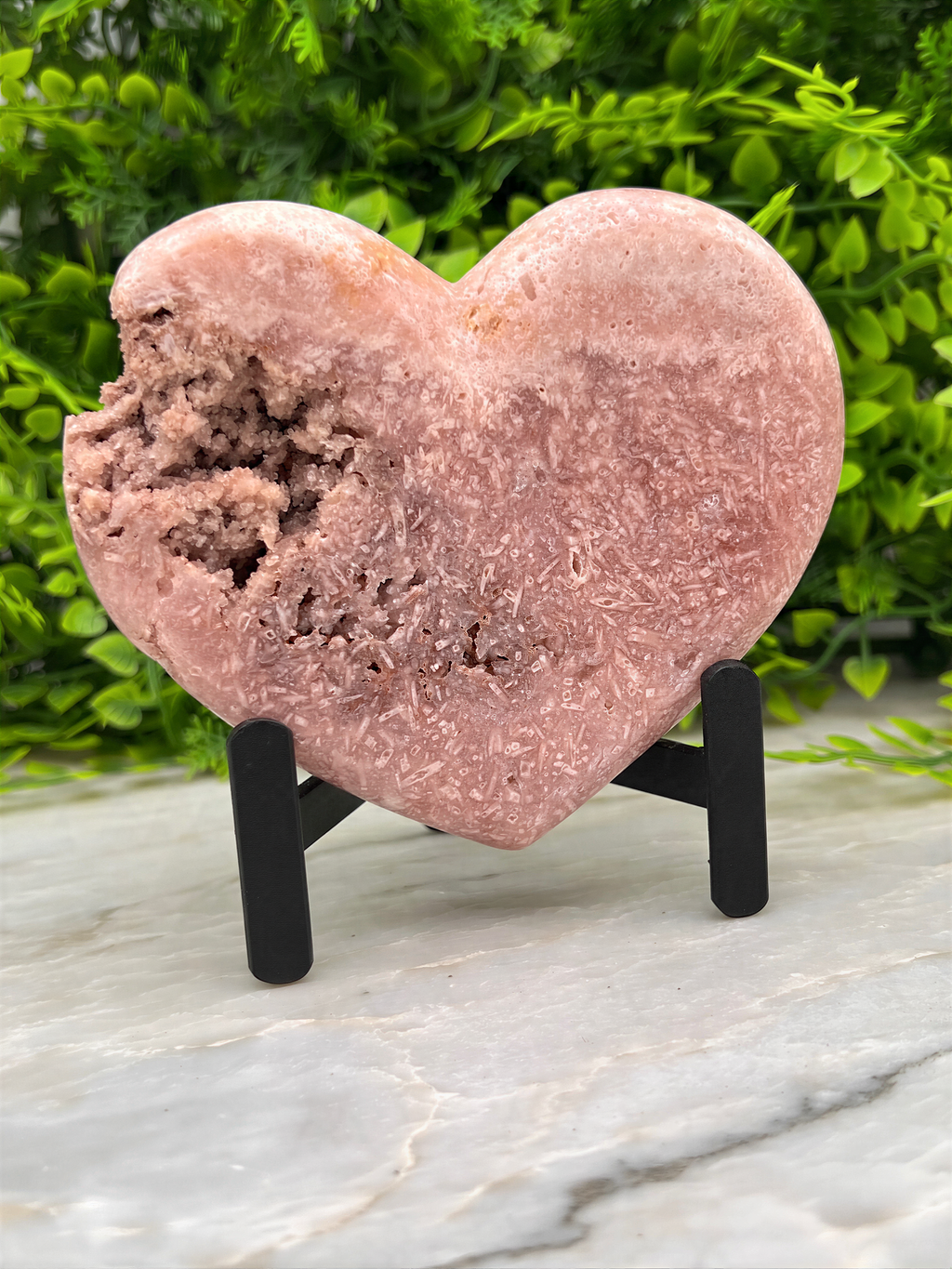 A heart-shaped pink crystal sits on a stand, surrounded by greenery.