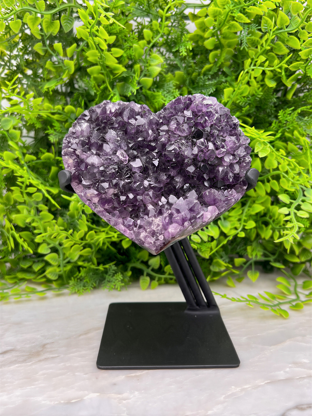 A heart-shaped amethyst crystal rests on a black stand, surrounded by lush green foliage.