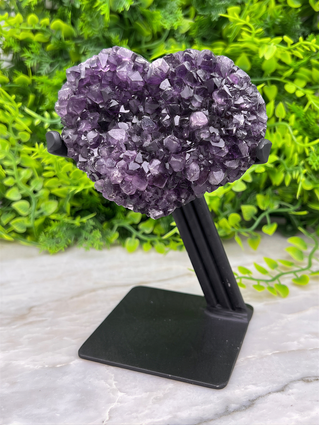 A heart-shaped crystal formation made of purple amethyst crystals is displayed on a black stand, surrounded by greenery.