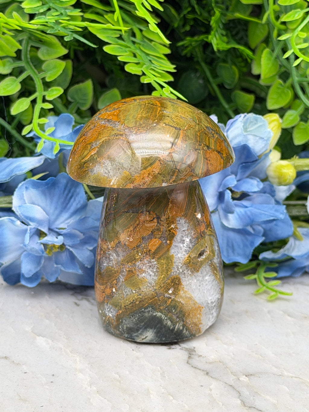 a close-up view of a natural stone, possibly a gemstone, resting on a white surface. The stone has a unique, textured pattern that resembles a mushroom or a similar organic shape. The stone is surrounded by a variety of green plants and blue flowers, creating a visually appealing contrast between the natural stone and the vibrant colors of the plants.