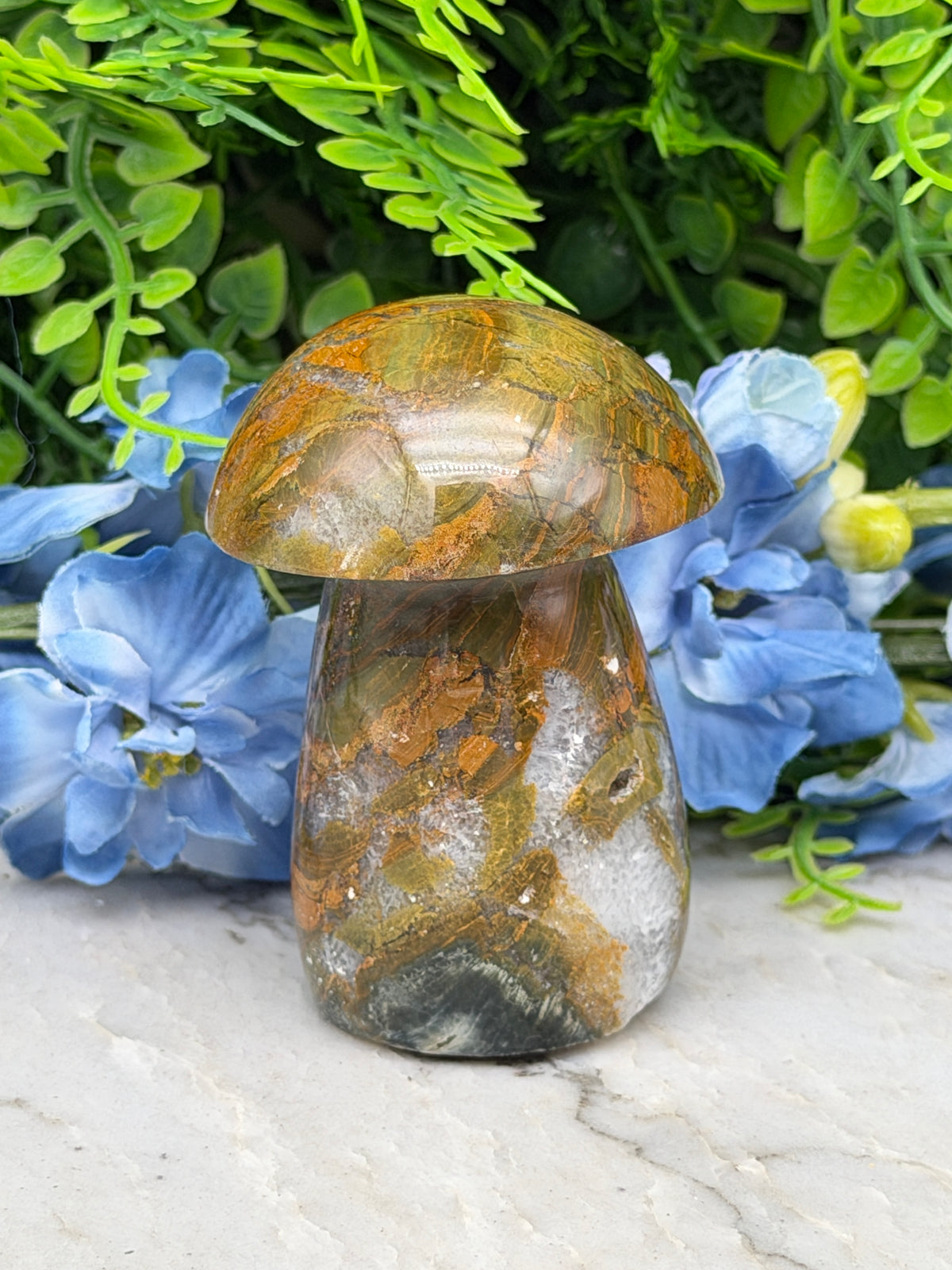 a close-up view of a natural stone, possibly a gemstone, resting on a white surface. The stone has a unique, textured pattern that resembles a mushroom or a similar organic shape. The stone is surrounded by a variety of green plants and blue flowers, creating a visually appealing contrast between the natural stone and the vibrant colors of the plants.