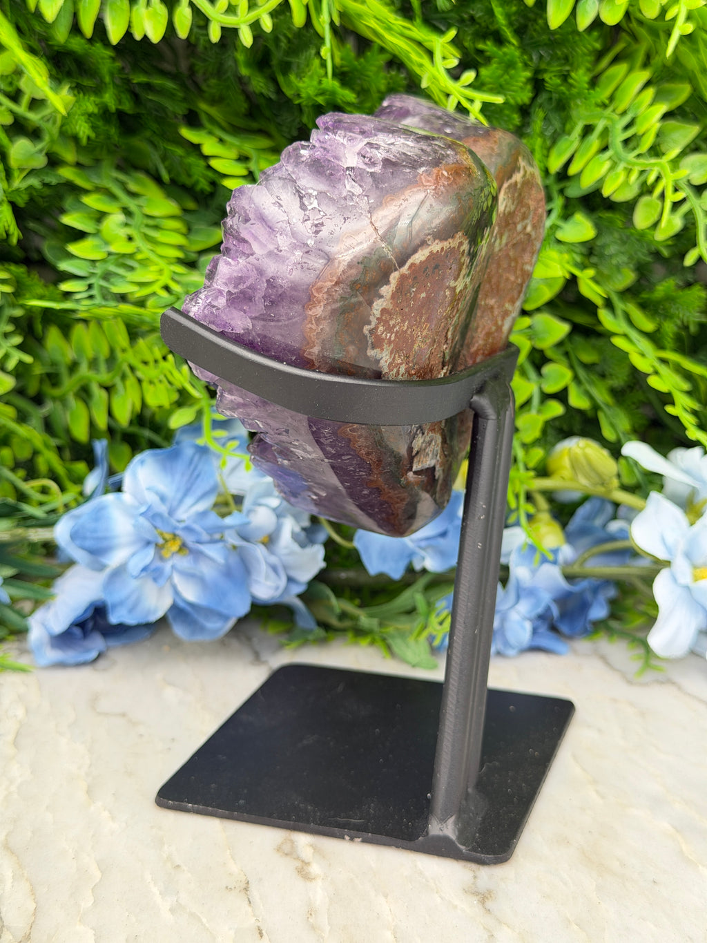 A large, multicolored rock is mounted on a black metal bracket, surrounded by a lush green plant and blue flowers.