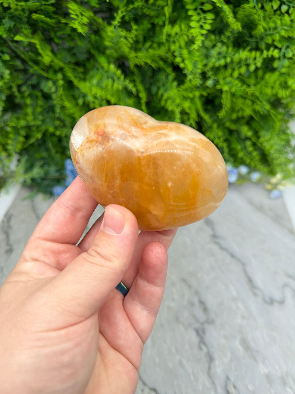 A hand holding a heart-shaped, orange-colored stone against a backdrop of green foliage.