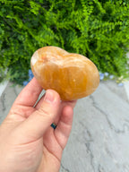 A hand holding a heart-shaped, orange-colored stone against a backdrop of green foliage.