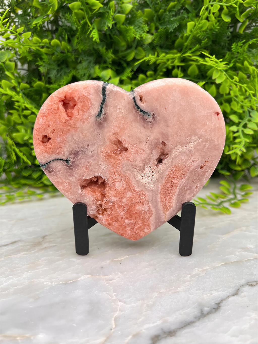 A pink geode-like rock formation is displayed on a stand, surrounded by greenery.