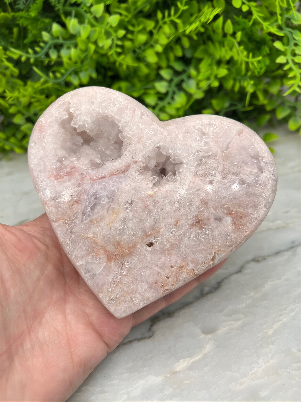 A hand is holding a heart-shaped geode with crystals inside, set against a backdrop of green foliage.