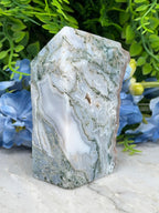 a close-up view of a jade crystal surrounded by lush green foliage and delicate blue flowers.