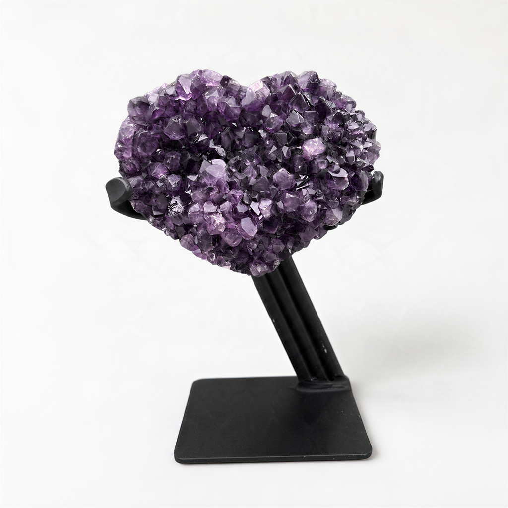 A heart-shaped cluster of purple amethyst crystals is displayed on a black stand against a white background.