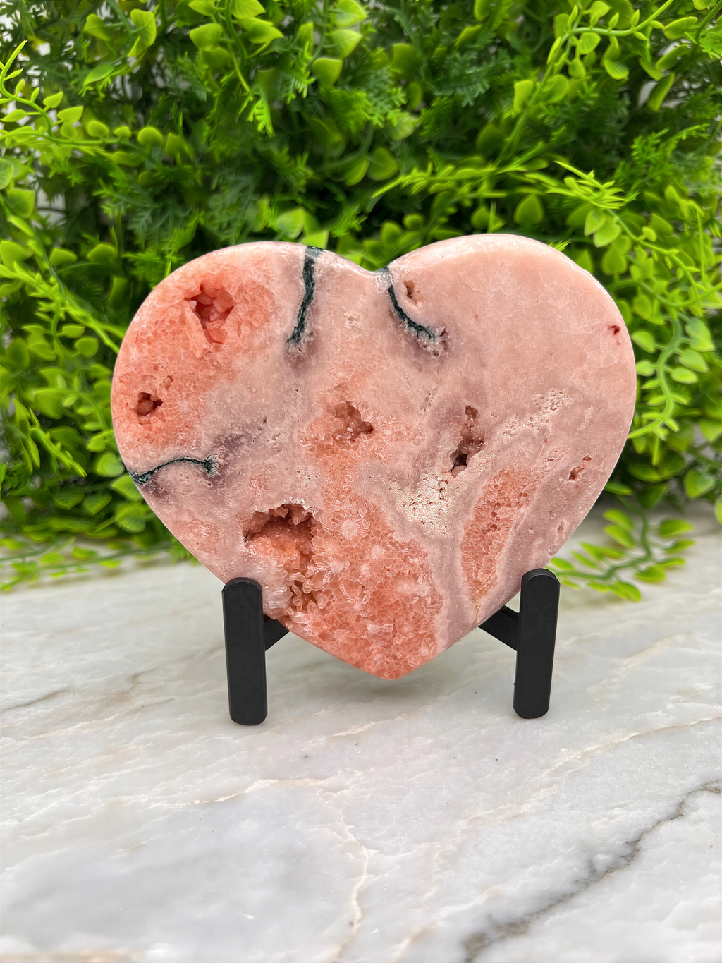 A pink geode-like rock formation is displayed on a stand, surrounded by greenery.