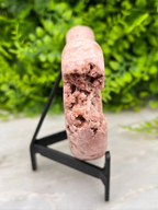 A pink crystal formation is displayed on a black stand, against a blurred green background.