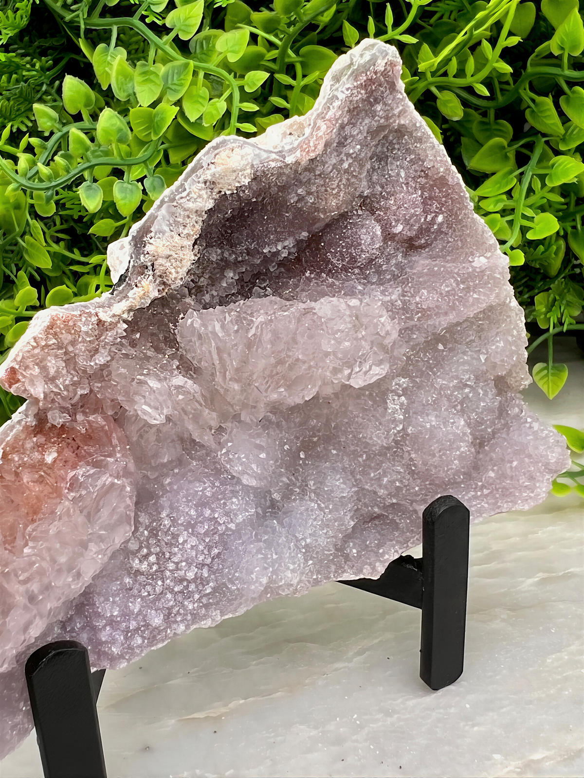 a large, irregularly shaped piece of pink crystal or rock, which appears to be a natural formation. The crystal is placed on a black stand, and there are some green plants visible in the background.