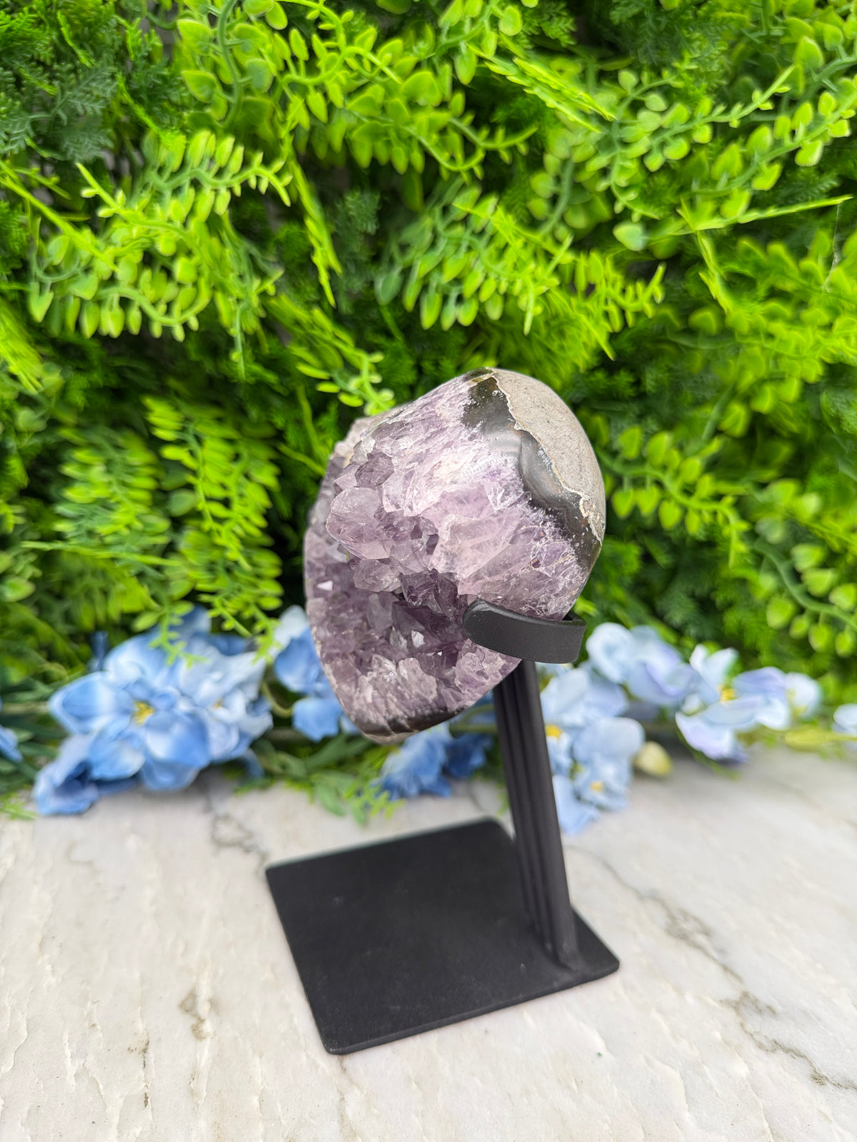 A close-up view of a purple crystal or gemstone, surrounded by lush green foliage and blue flowers, mounted on a black stand or display.