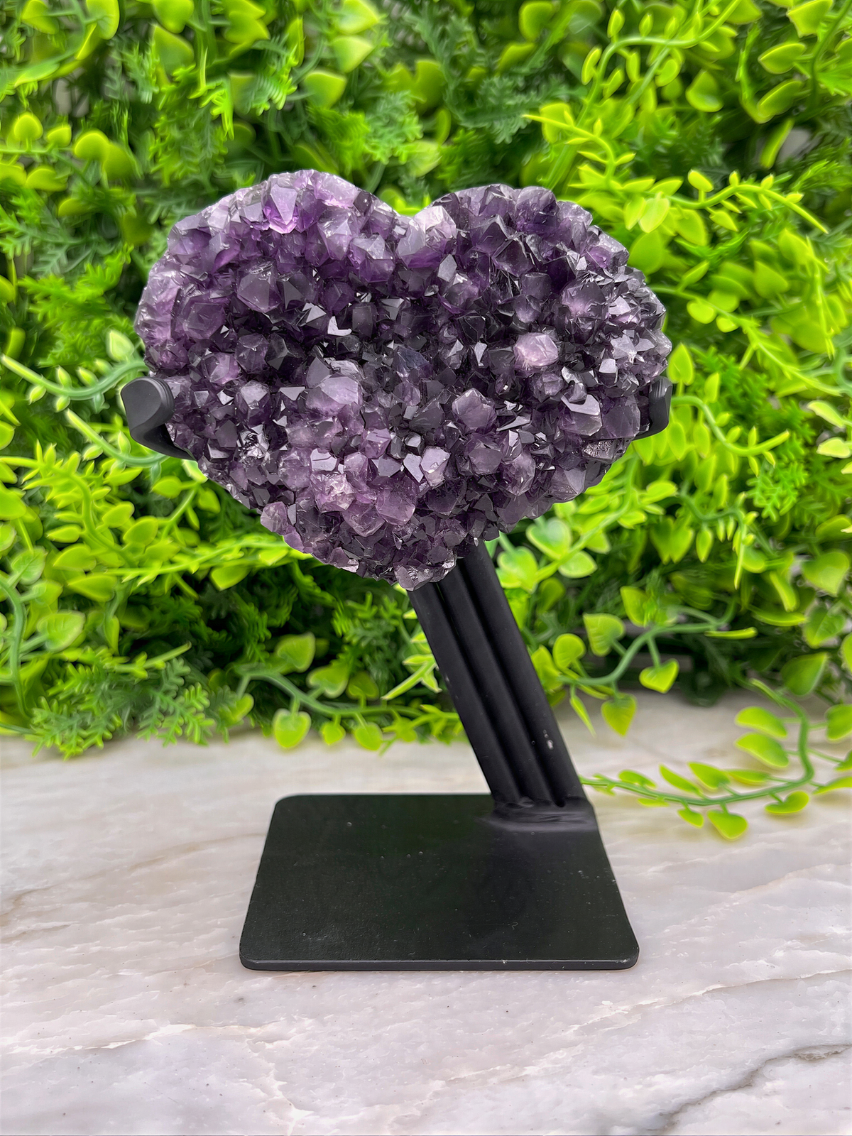 A heart-shaped crystal formation made of purple amethyst rests on a black stand, surrounded by lush green foliage.