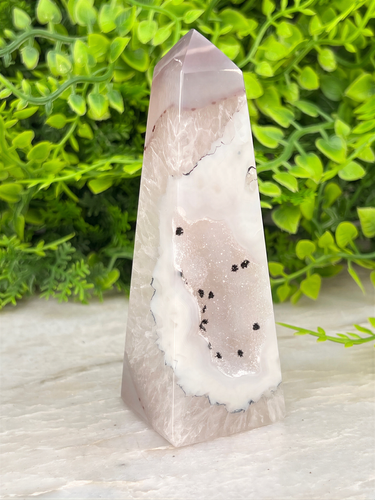 A pink and white geode-like crystal rests on a white surface, surrounded by lush green foliage.