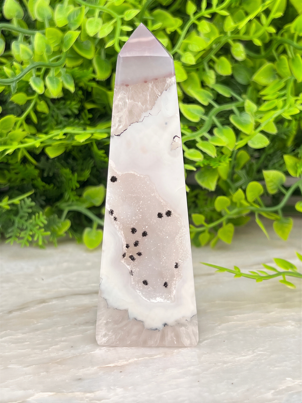 A pink and white crystal formation with black spots is displayed against a backdrop of green foliage.
