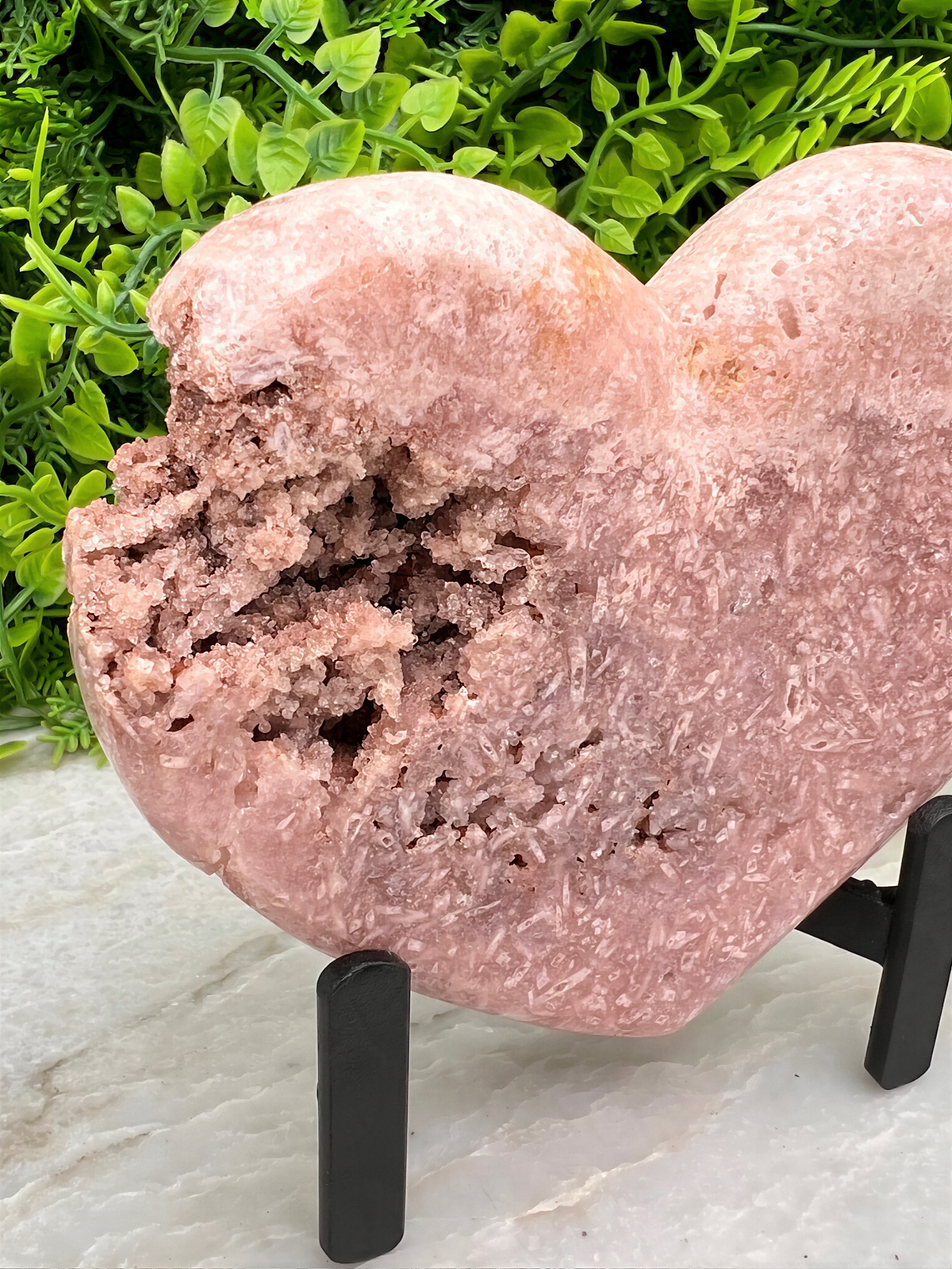 A pink crystal heart-shaped object is displayed on a stand, surrounded by greenery.