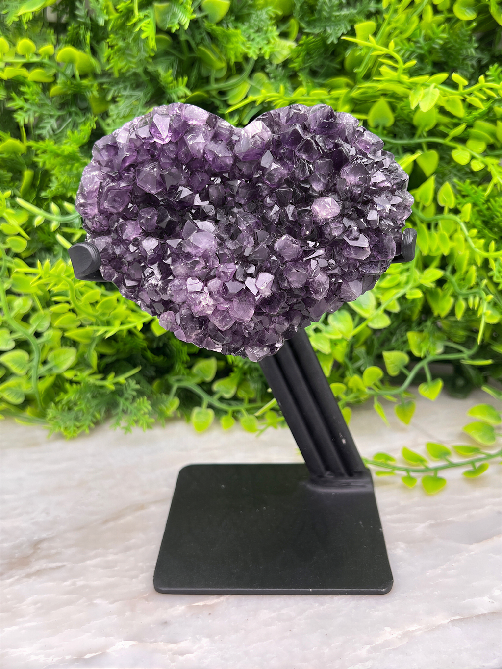 A heart-shaped cluster of purple amethyst crystals is displayed on a black stand, set against a backdrop of lush green foliage.