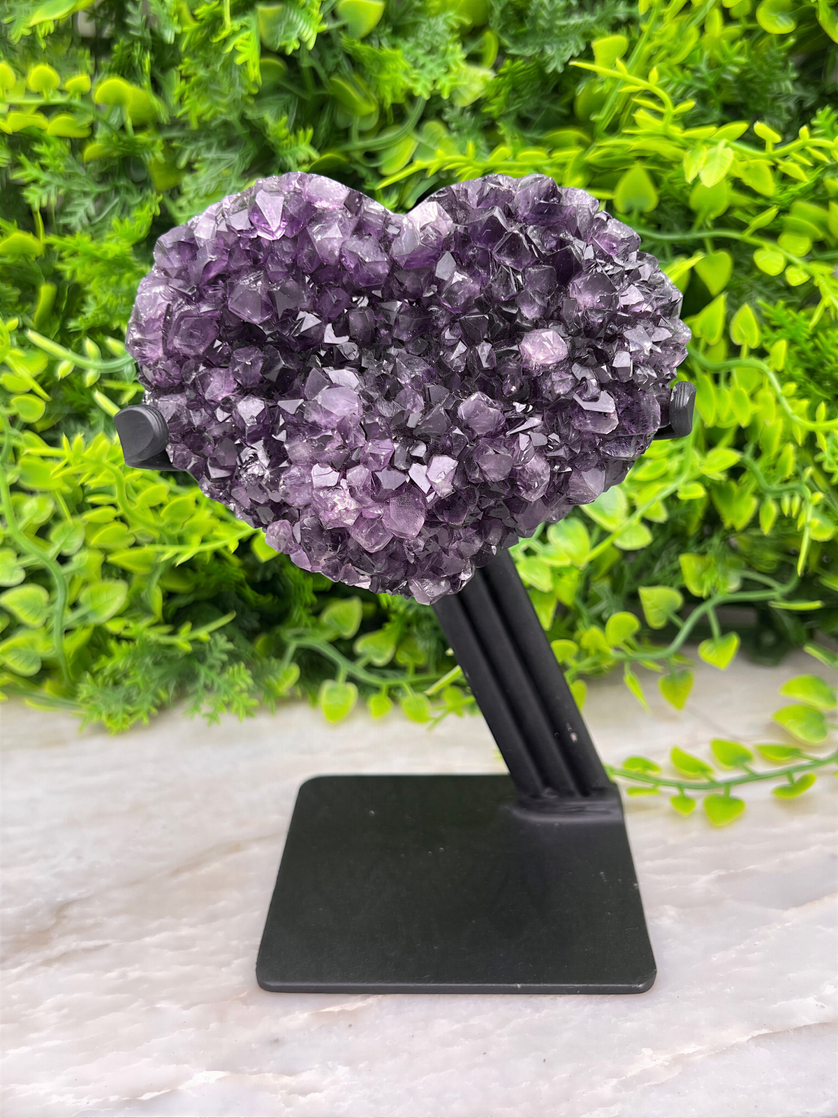 A heart-shaped cluster of purple amethyst crystals is displayed on a black stand, set against a backdrop of lush green foliage.