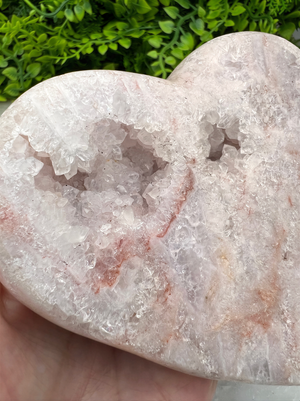 A hand holds a heart-shaped crystal formation made of pink and white minerals, with a green plant in the background.