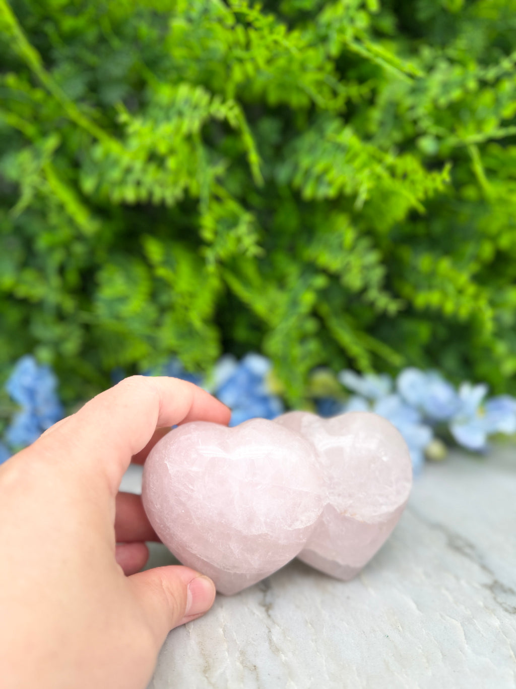 A hand is holding two pink crystal hearts in front of a lush green background with blue flowers.