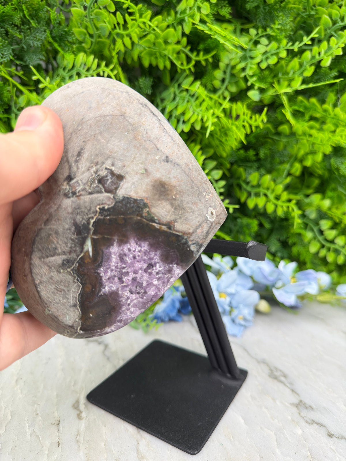 A hand holding a rock with a purple gemstone embedded in it, set against a backdrop of green foliage and blue flowers.