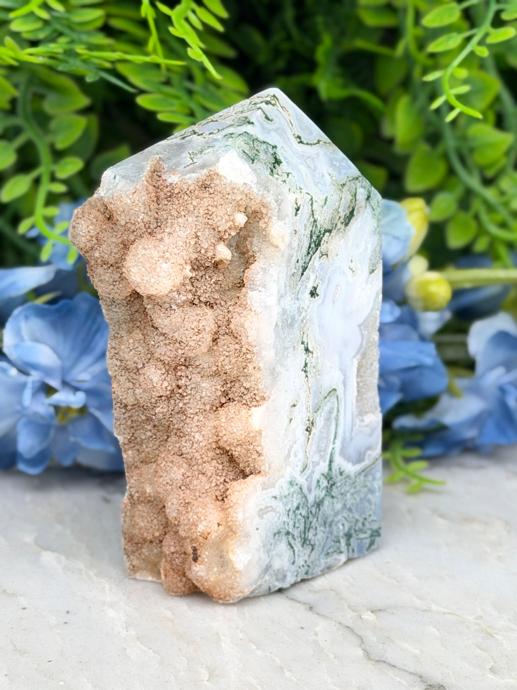a close-up of a jade crystal with a mix of white and brown hues, set against a backdrop of lush green foliage and delicate blue flowers.