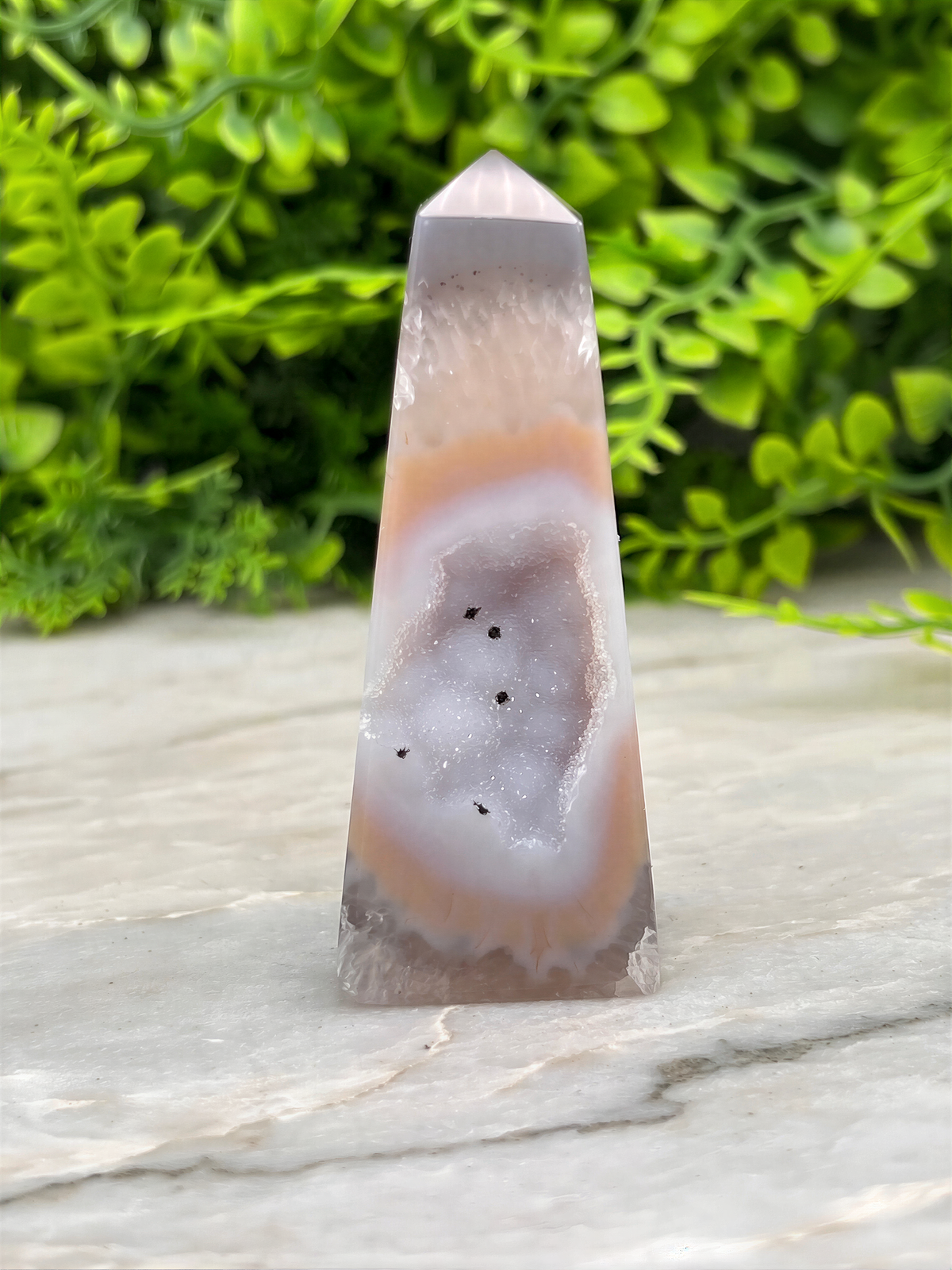A clear geode with a unique rock formation sits on a marble surface, surrounded by lush green foliage.