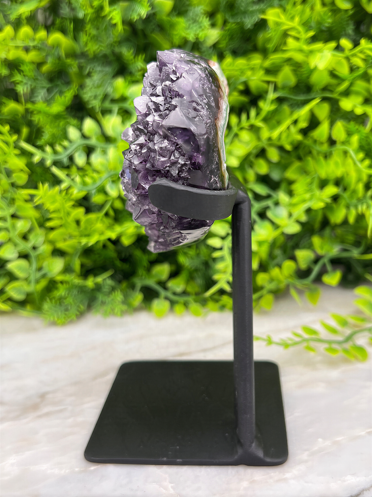 A large, purple amethyst crystal is displayed on a black metal stand, surrounded by lush green foliage.