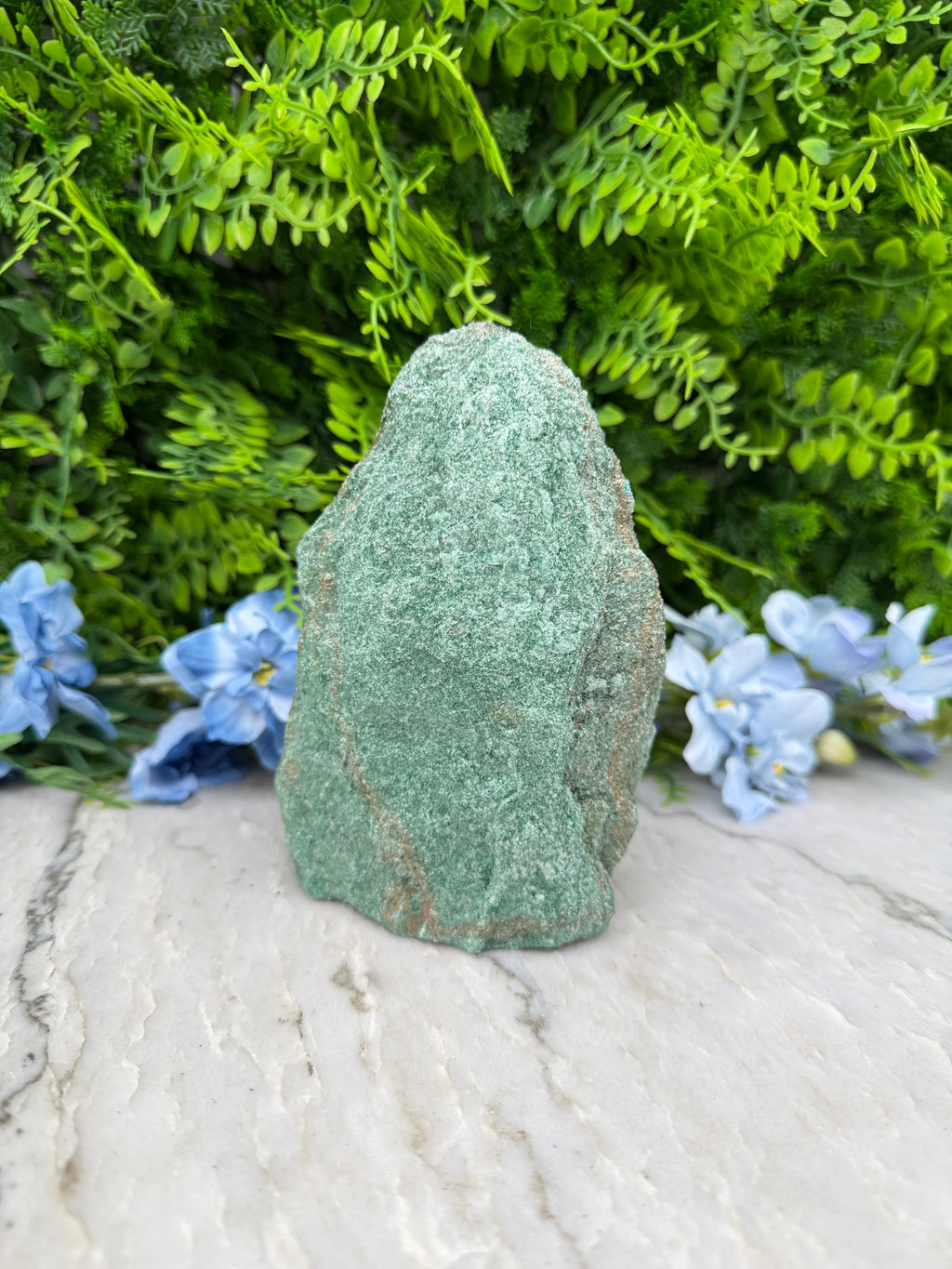 A large, rough-textured green rock rests on a white surface, surrounded by lush green foliage and delicate blue flowers.