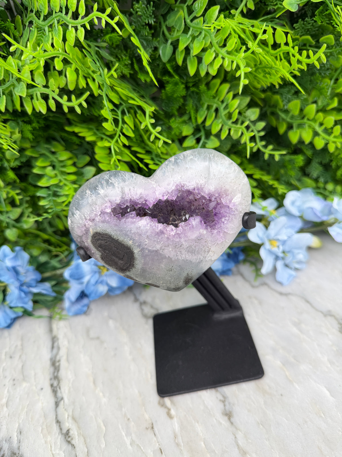 A heart-shaped amethyst geode is displayed on a white marble surface, surrounded by lush green foliage and blue flowers.