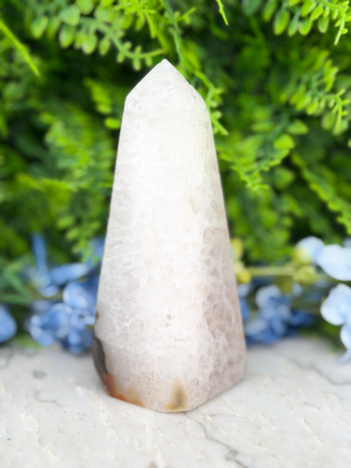 A white, triangular crystal sits on a white surface, surrounded by lush green foliage.