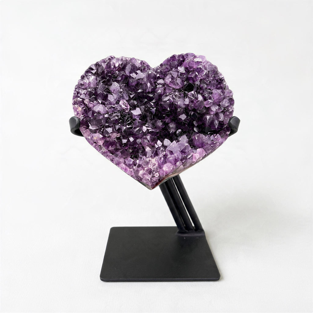A heart-shaped crystal formation made of purple crystals is displayed on a black stand against a white background.