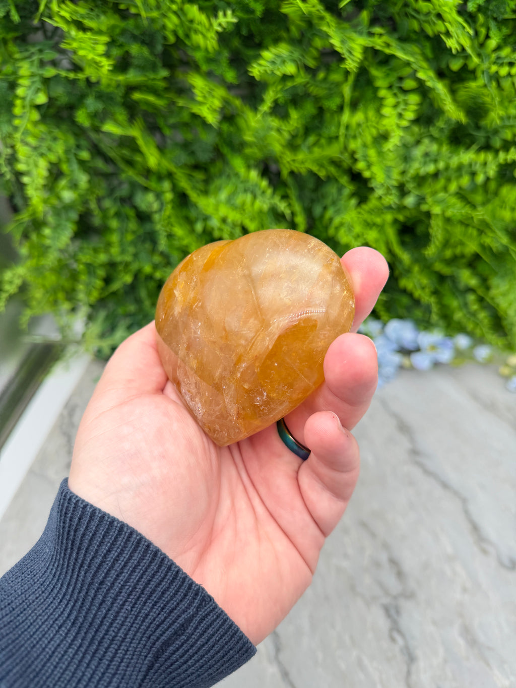 A hand holding a yellowish-brown crystal or gemstone against a backdrop of green foliage.