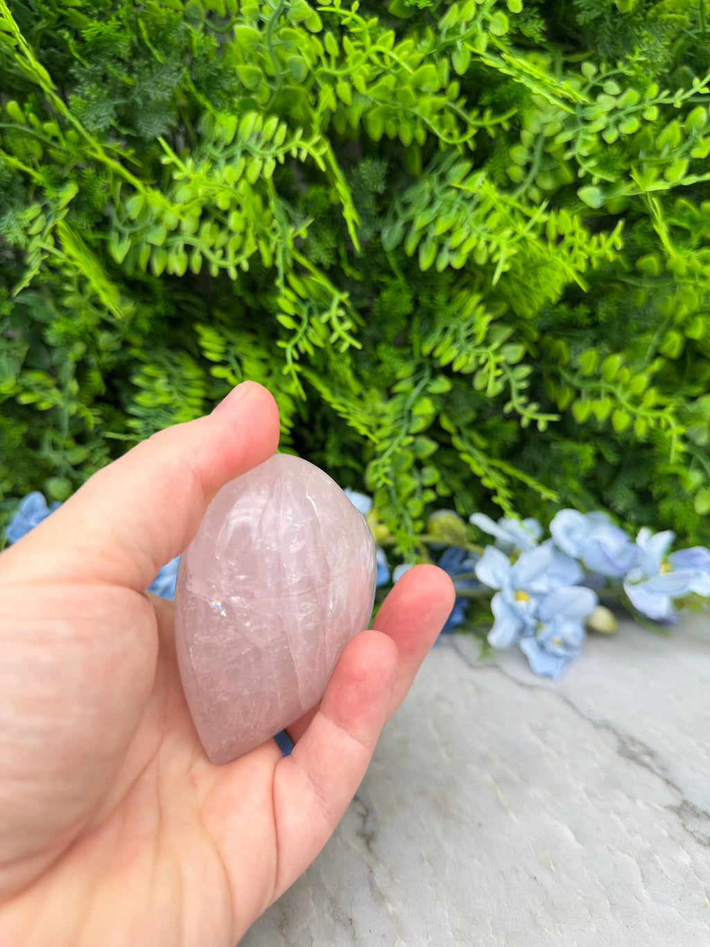 A hand holding a pink crystal against a backdrop of green foliage and blue flowers.
