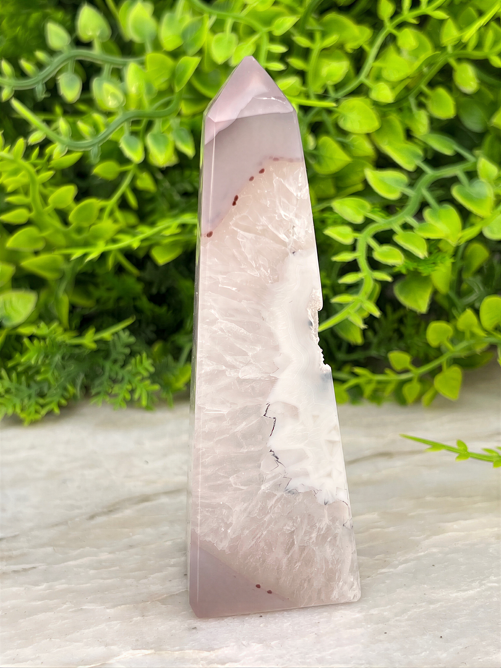 A pink jade crystal with a unique pattern rests on a white surface, surrounded by lush green foliage.