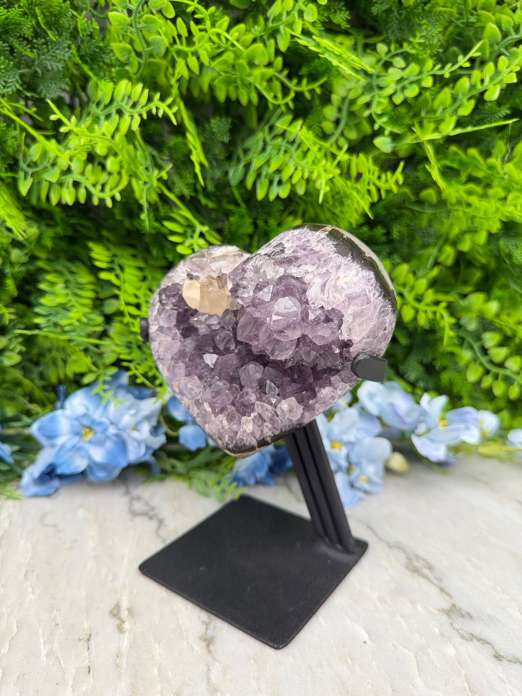 A heart-shaped crystal formation is displayed on a stand, surrounded by lush green foliage and blue flowers.
