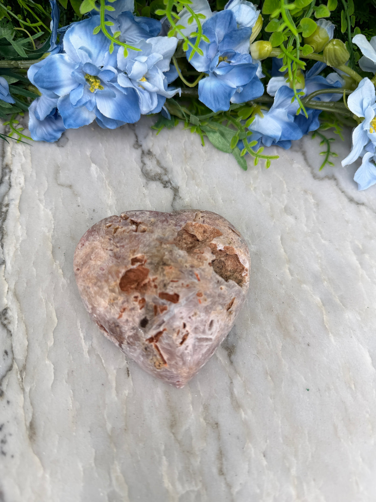 a heart-shaped rock with a brown and white speckled surface, placed on a white marble surface next to a cluster of blue flowers.