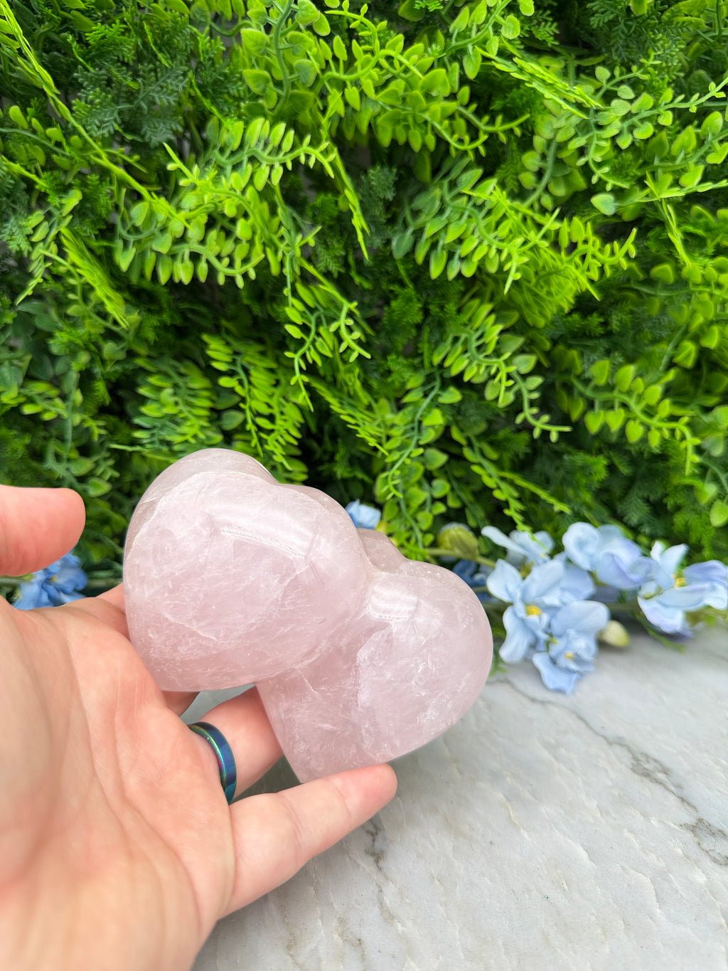A hand holding two heart-shaped crystals against a backdrop of lush green foliage and delicate blue flowers.