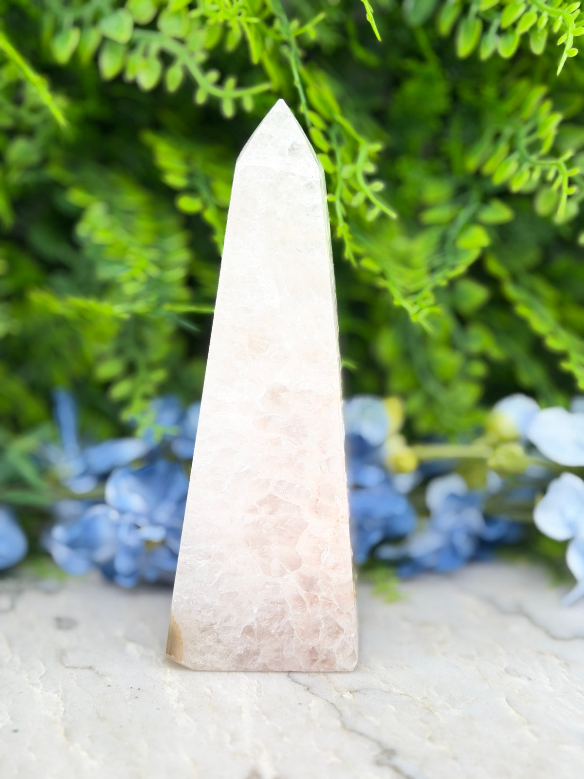 A white, triangular crystal formation is prominently displayed in the center of the image, surrounded by a blurred background of green foliage.