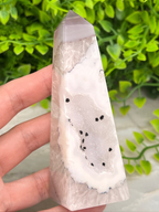 A hand is holding a clear crystal with a unique, irregular shape and a pattern of black spots on its surface, set against a backdrop of green foliage.