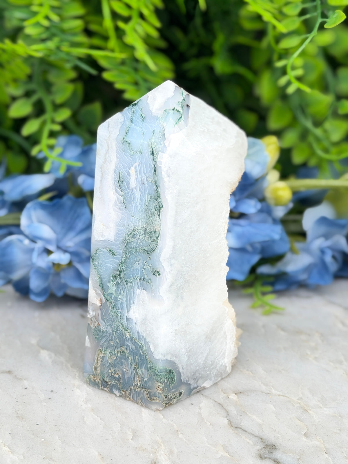 a white and green crystal with a jagged edge, placed on a white surface. The crystal is surrounded by blue flowers, creating a visually appealing contrast.