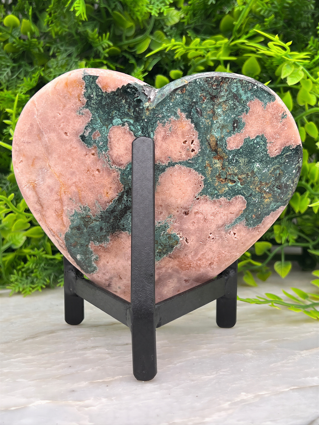 A heart-shaped rock with a green and pink color pattern sits on a black stand, surrounded by greenery.