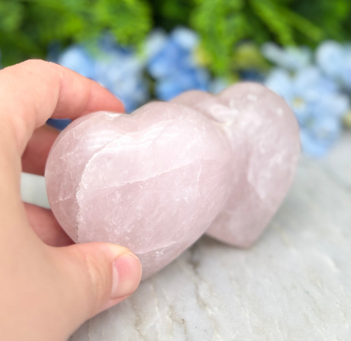 A hand is holding two pink crystal hearts against a backdrop of lush green foliage and blue flowers.