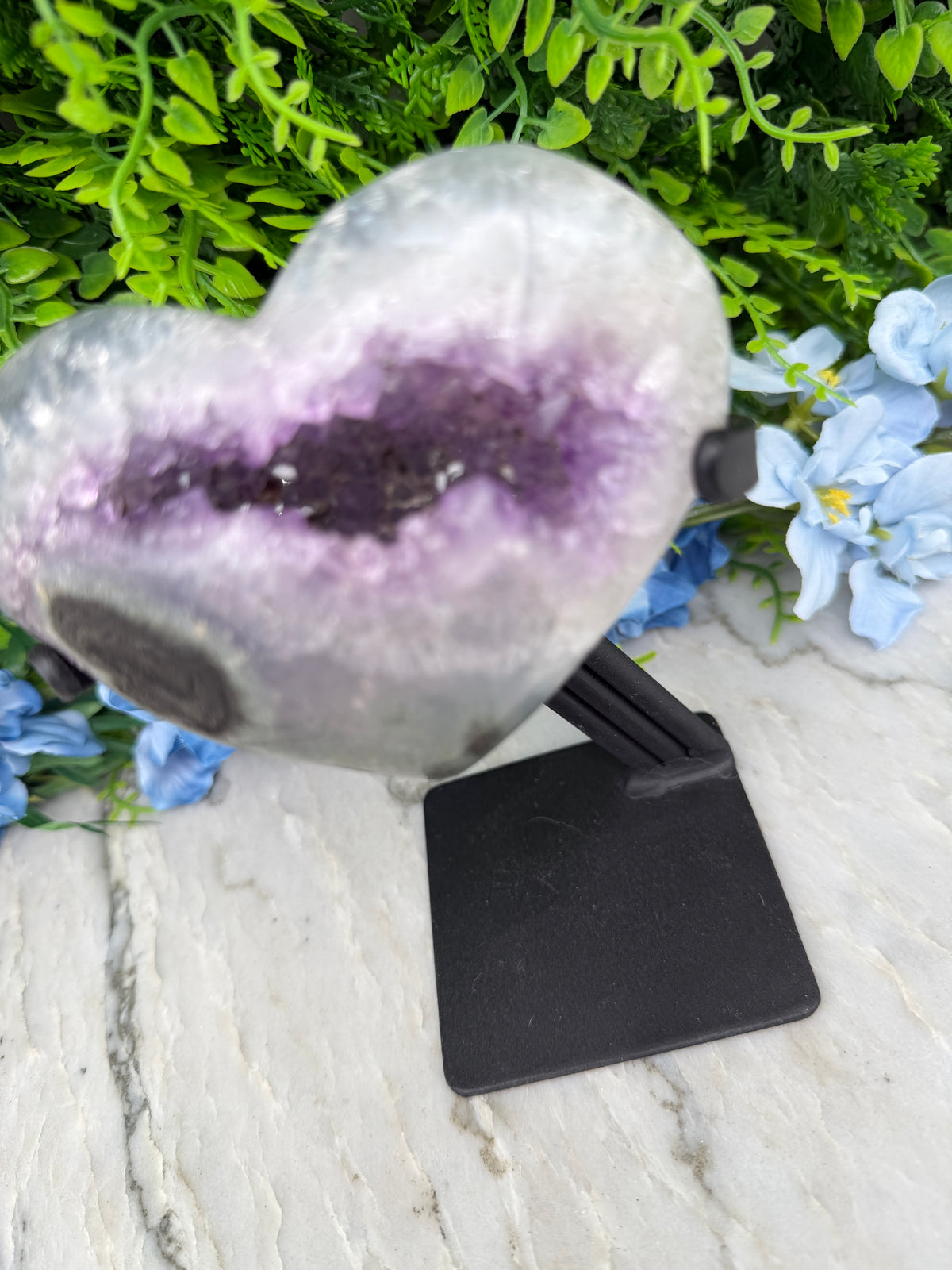a large purple amethyst geode with a black stand, set against a backdrop of green foliage and blue flowers.